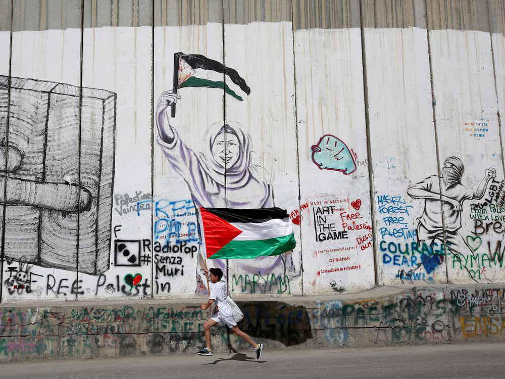 Korkea apartheidmuuri, jossa on graffiteja, ja edessä juoksee pieni lapsi kädessään Palestiinan lippu.